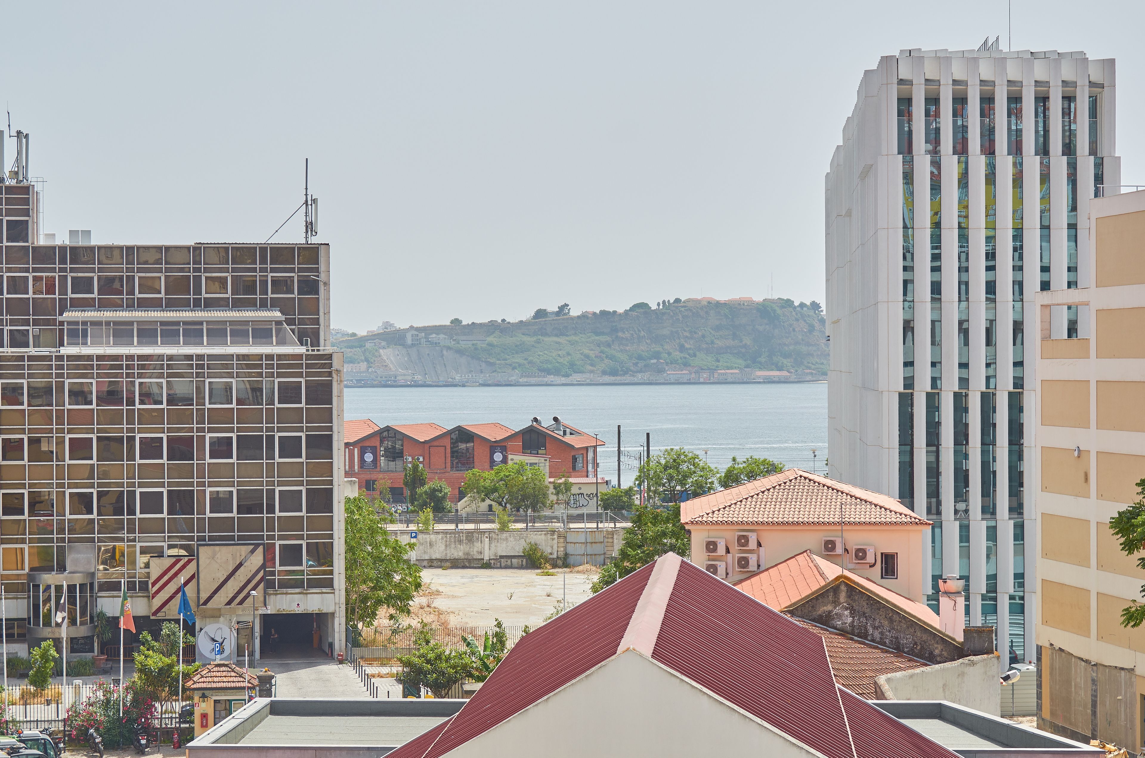 A modern building with a glass facade, a red-roofed building in the foreground, and a body of water in the background.
