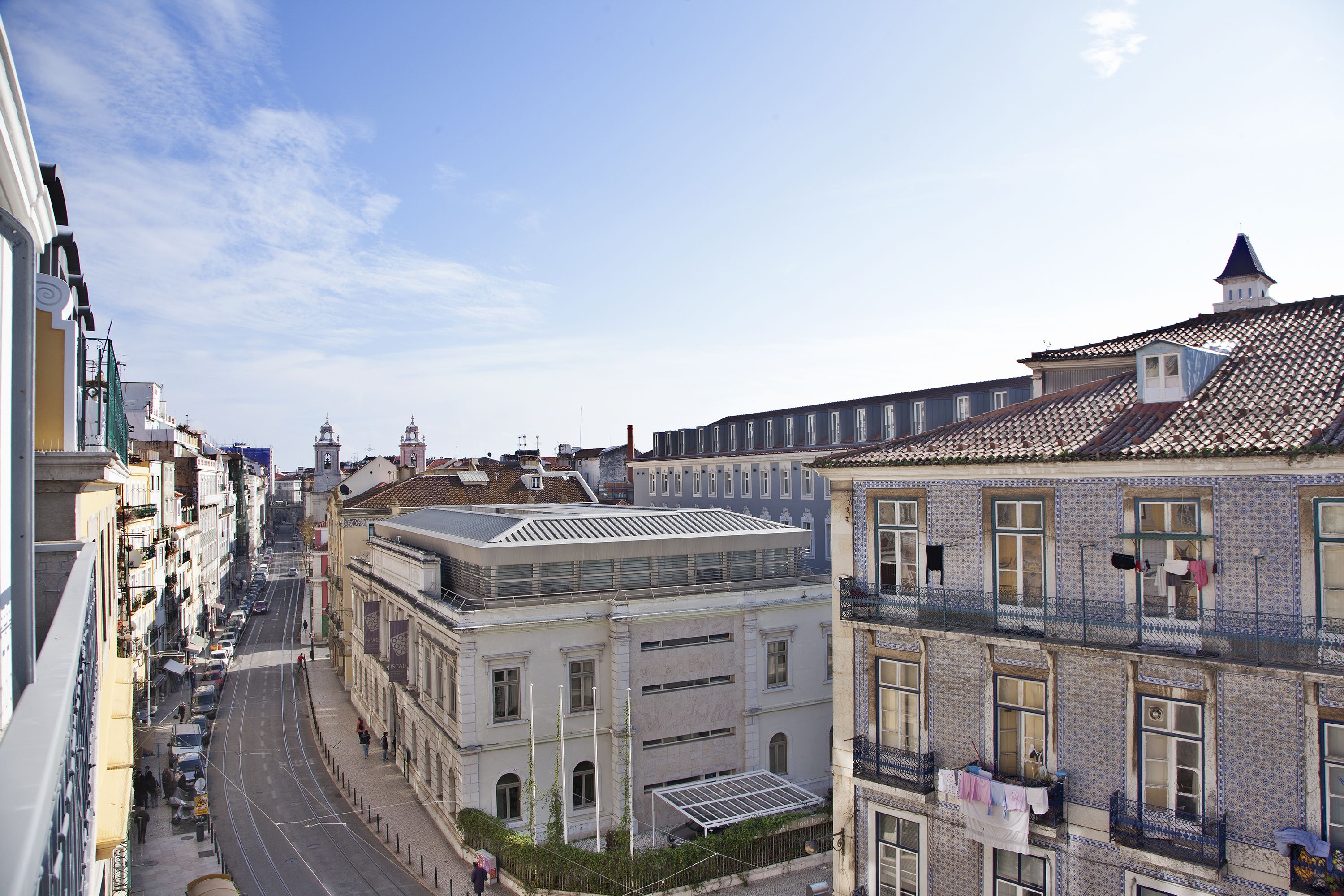 A street view of a city with buildings, including a prominent building with a flat roof and a balcony with clothes hanging.