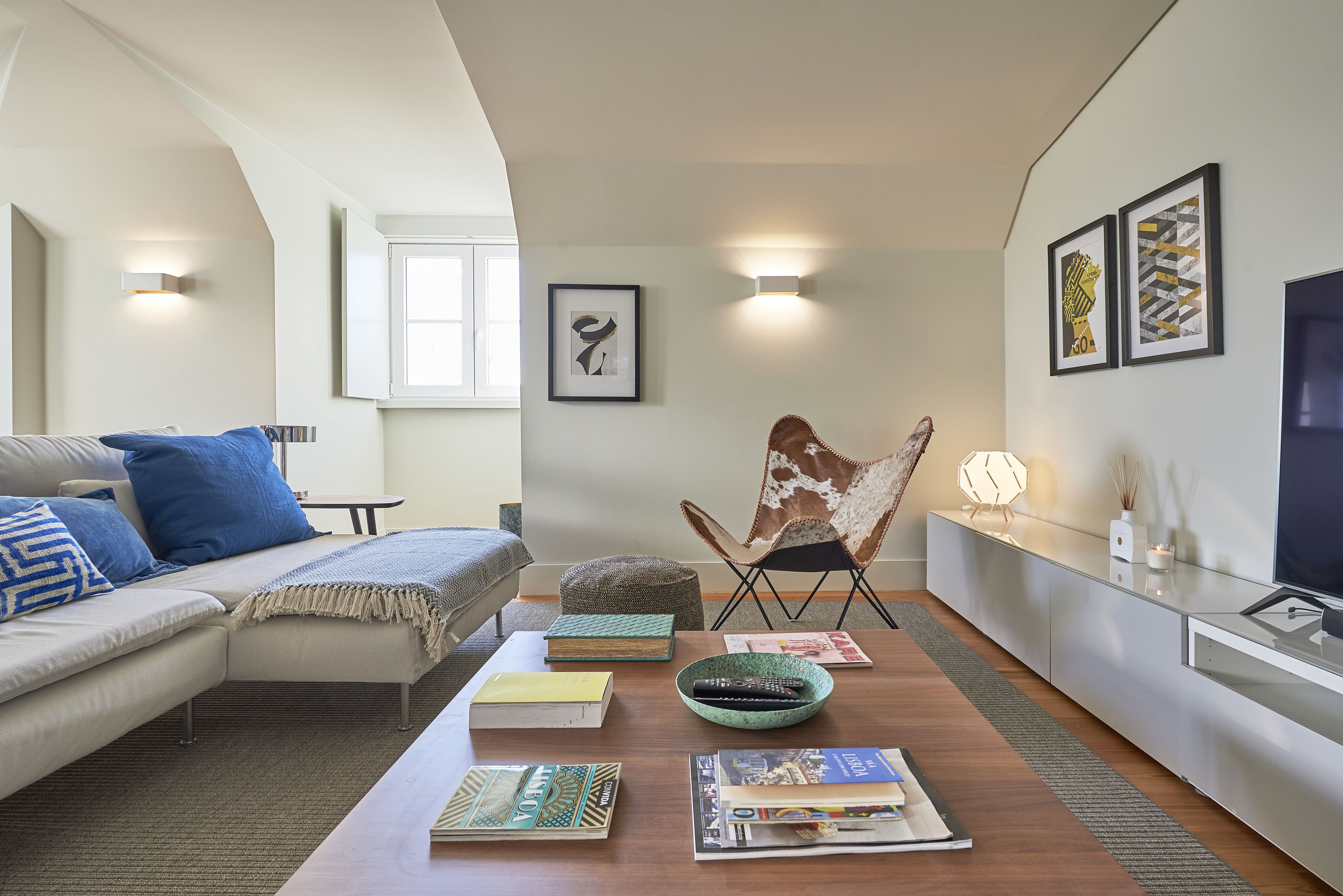 Ein modernes Wohnzimmer mit einer Couch, einem Couchtisch mit Büchern und Fernbedienungen, einem Zierstuhl und Wandbildern.