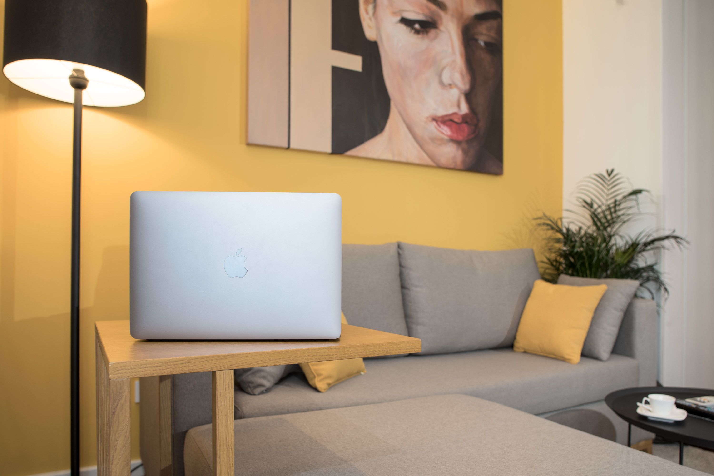 A laptop on a wooden table, a grey sofa with yellow cushions, a black floor lamp, and a small potted plant.