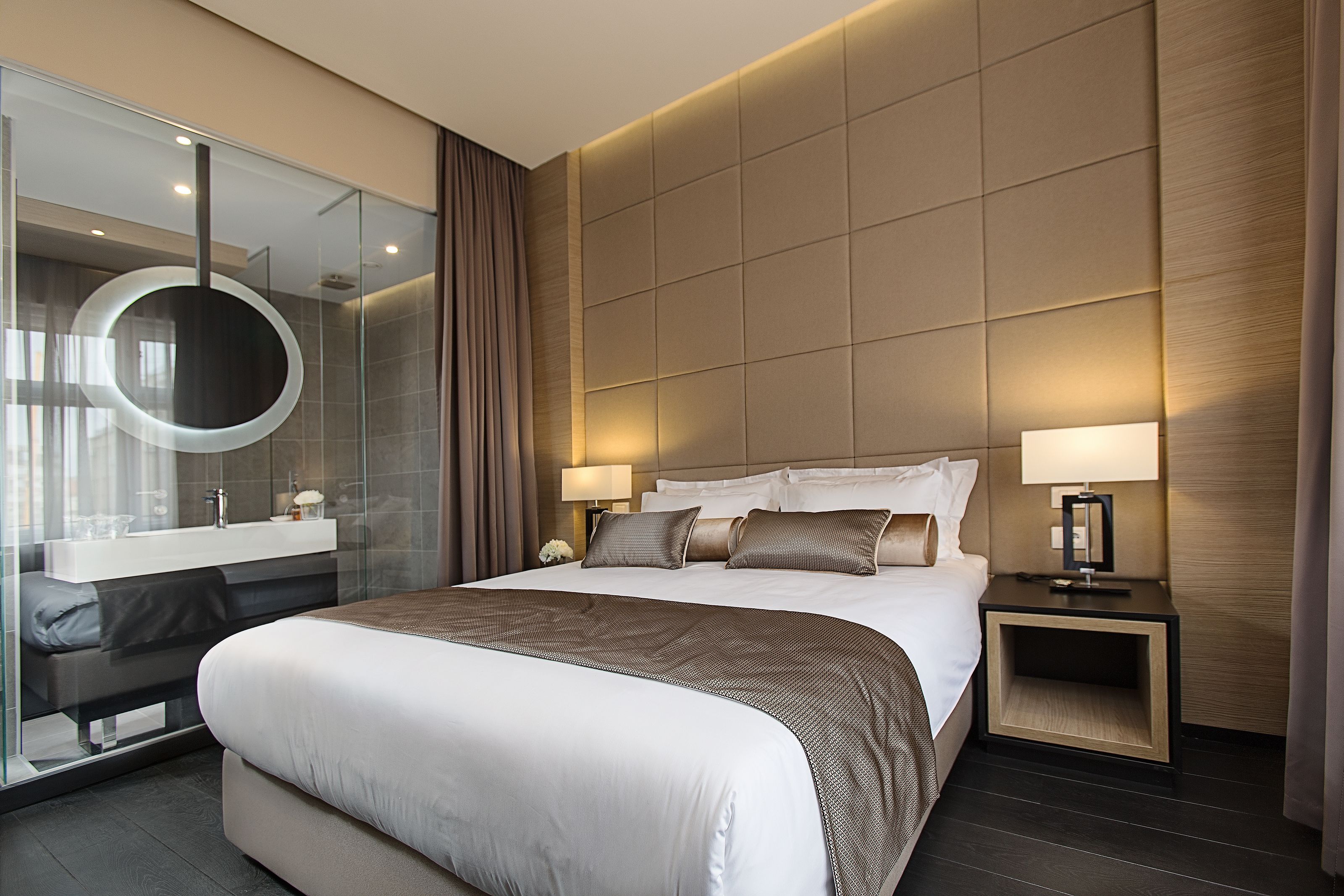 A modern hotel room with a large bed, bedside tables, a round mirror, and a glass partition.