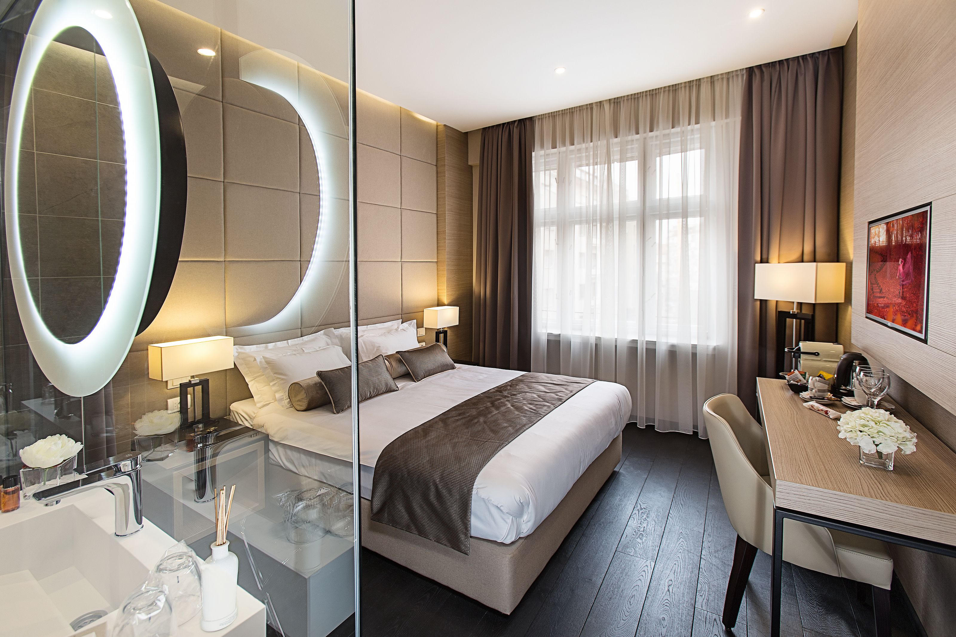 A modern hotel room with a large bed, a glass-enclosed bathroom, a dining area, and a large mirror.