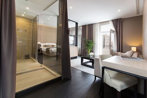 A modern hotel room with a glass shower enclosure, a bed with white linens, a dining area with a table and chairs, and a view of the city through large windows.