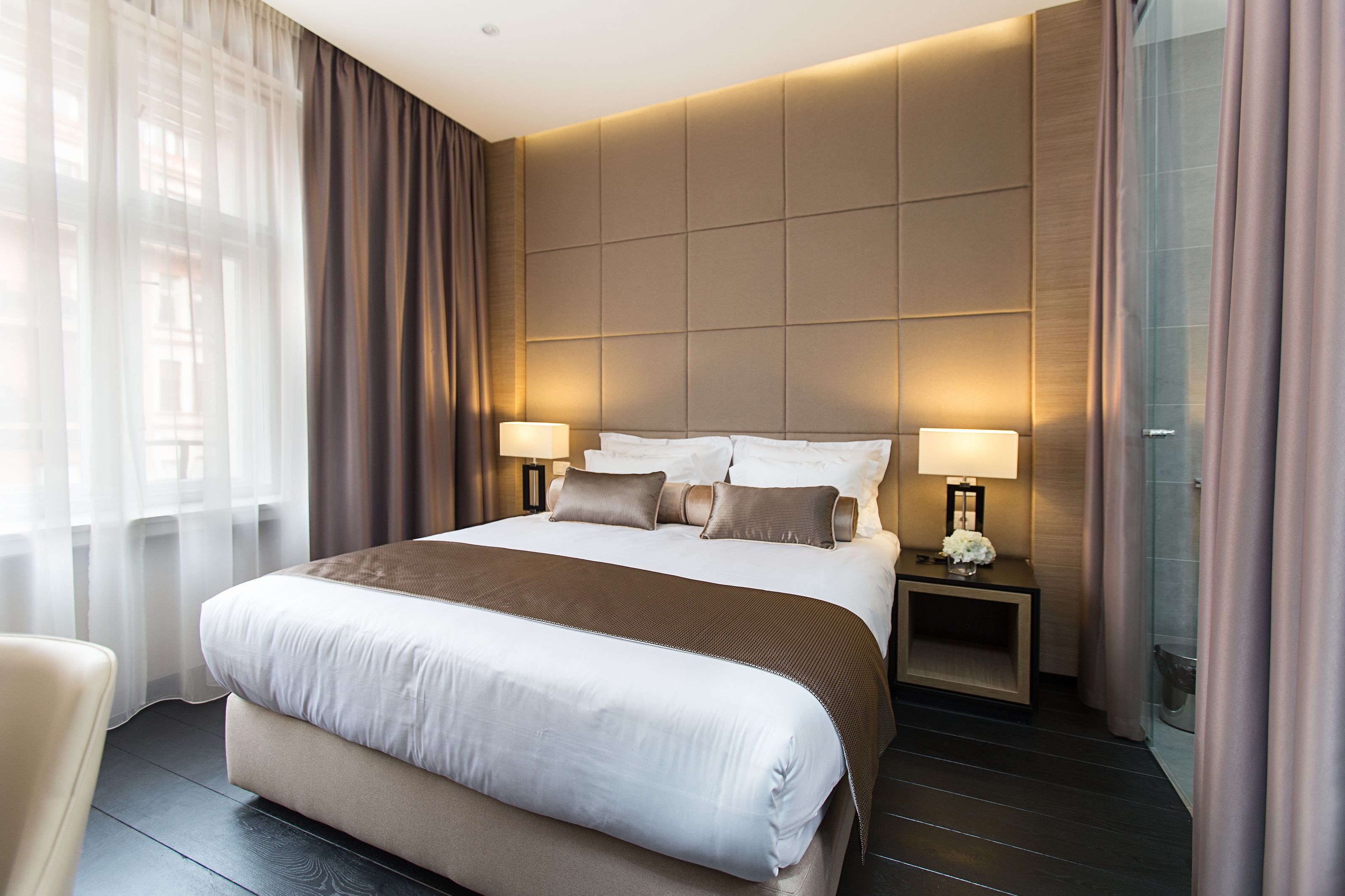 A modern hotel room with a large bed, two bedside lamps, a small table with a vase, and a view of the city through sheer curtains.