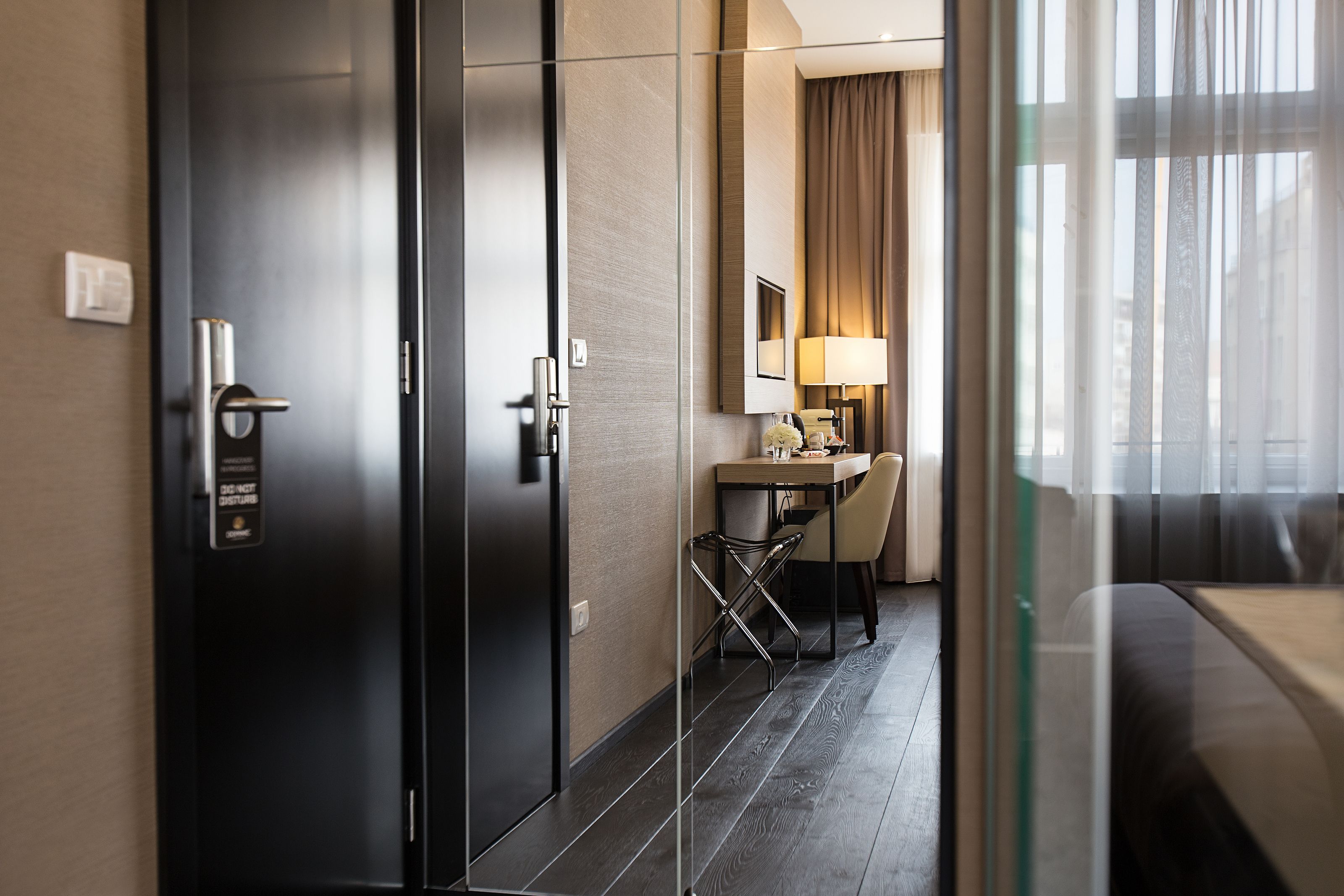 Ein Hotelzimmer mit einer dunklen Holztür, einem Schreibtisch mit Stuhl, einer Lampe und Blick durch eine Glasscheibe.