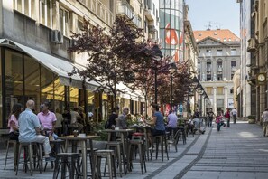 An outdoor café with tables and chairs, surrounded by buildings with large windows and a tree in the center. - Dominic & Smart Luxury Suites Republic Square (Belgrade)