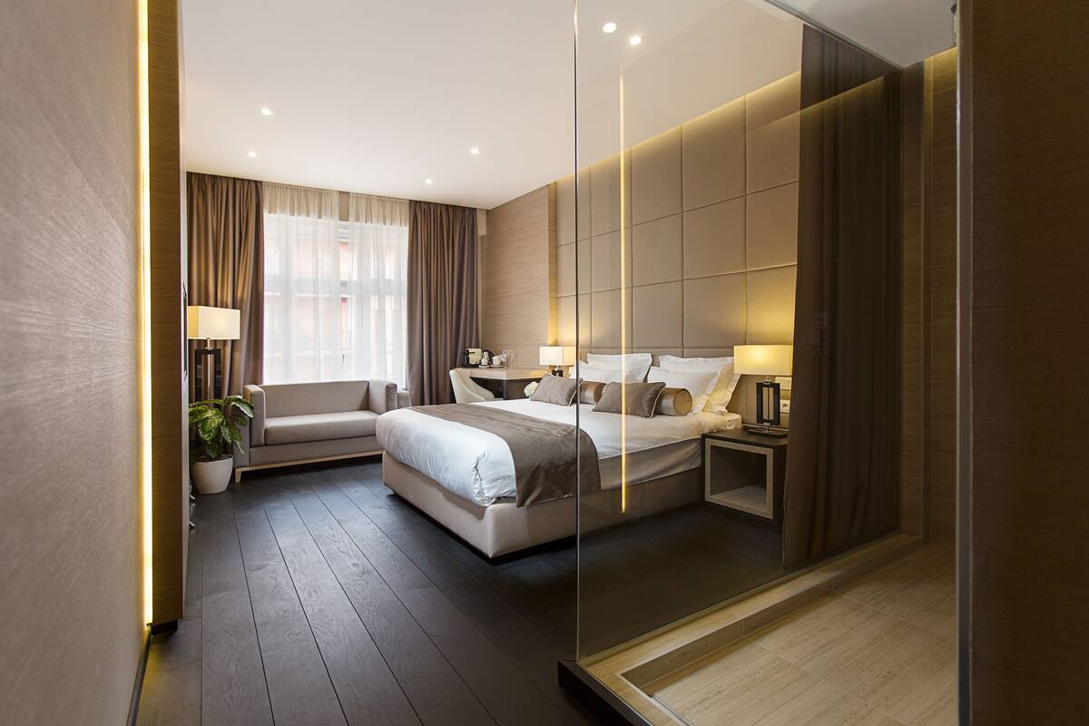 A modern hotel room with a large bed, a sofa, a bedside table, and a potted plant.