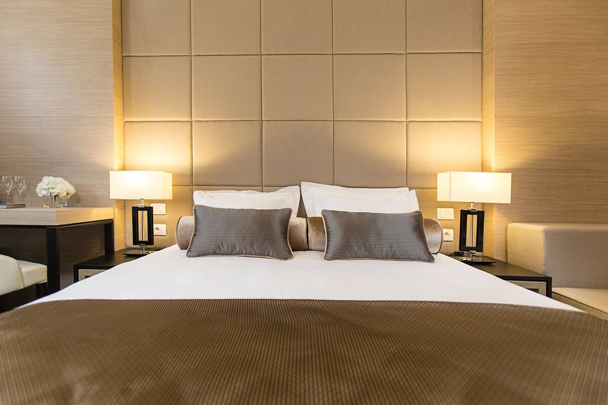 A hotel room with a large bed, two bedside tables with lamps, a small desk with a chair, and a sofa.