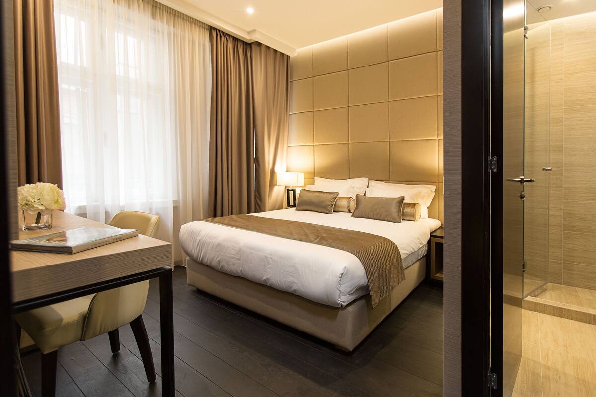 A modern hotel room with a large bed, a desk with a laptop, and a glass shower enclosure.