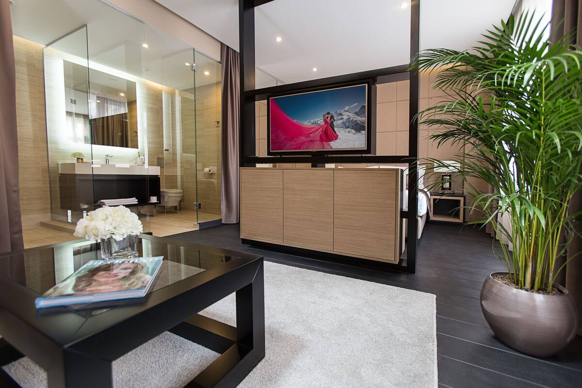 A modern hotel room with a glass shower enclosure, a wooden vanity, a large flat-screen TV, and a potted plant.