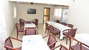 Un espace repas comprenant plusieurs tables et chaises, un téléviseur fixé au mur et un réfrigérateur orné d’un logo.