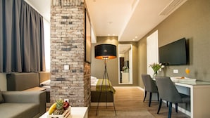 A modern living room with a brick accent wall, a sofa, a coffee table with fruit, a floor lamp, and a TV mounted on the wall. - Hotel Belgrade Inn (Belgrade)