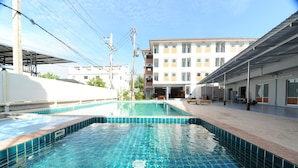 Une piscine dont les bords sont recouverts de carreaux, un espace abrité avec des bancs, et un immeuble à plusieurs étages en arrière-plan.