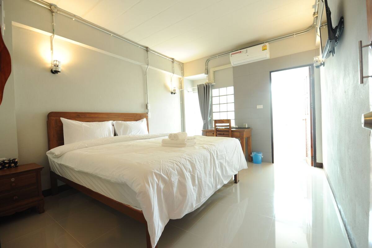 A clean, well-lit hotel room with a large bed, bedside tables, a wooden nightstand, a small desk, and a chair.