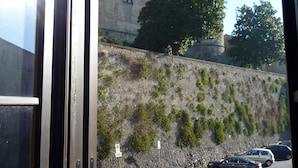 Vista a través de una ventana que muestra un estacionamiento con coches, un muro de piedra y un edificio con aspecto de castillo al fondo.