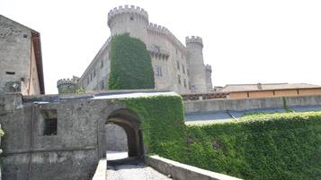 Un castello storico in pietra con una torre centrale, un arco d'ingresso e un muro circondato da vegetazione.