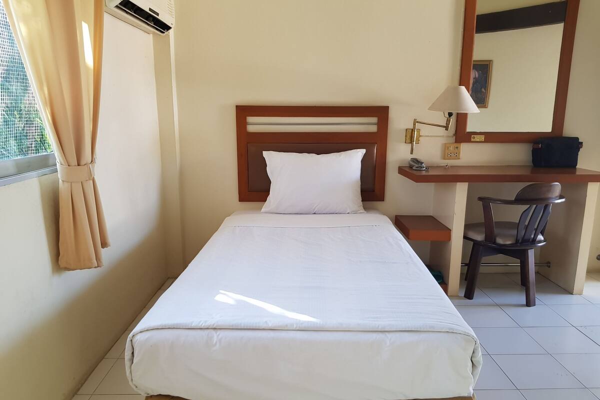 Un lit simple avec une literie blanche, une tête de lit en bois, un bureau avec une chaise, une lampe et un miroir.