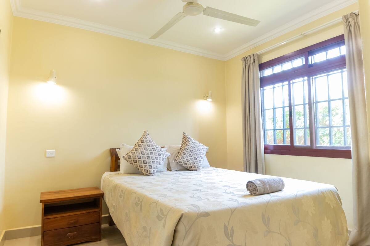 Une chambre à coucher avec un lit, une table de chevet, un ventilateur de plafond et une fenêtre avec des rideaux.