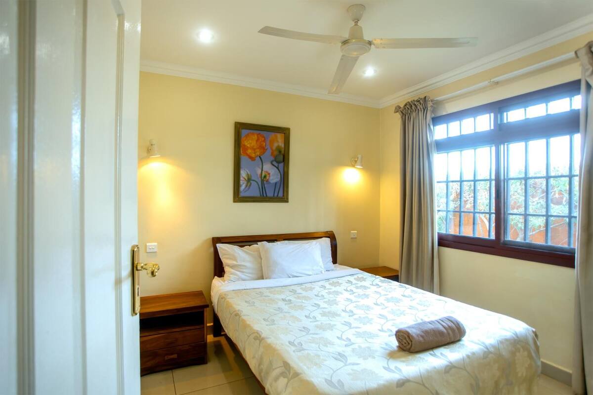 Une chambre à coucher avec un lit, un ventilateur de plafond, une fenêtre avec des rideaux et un tableau accroché au mur.