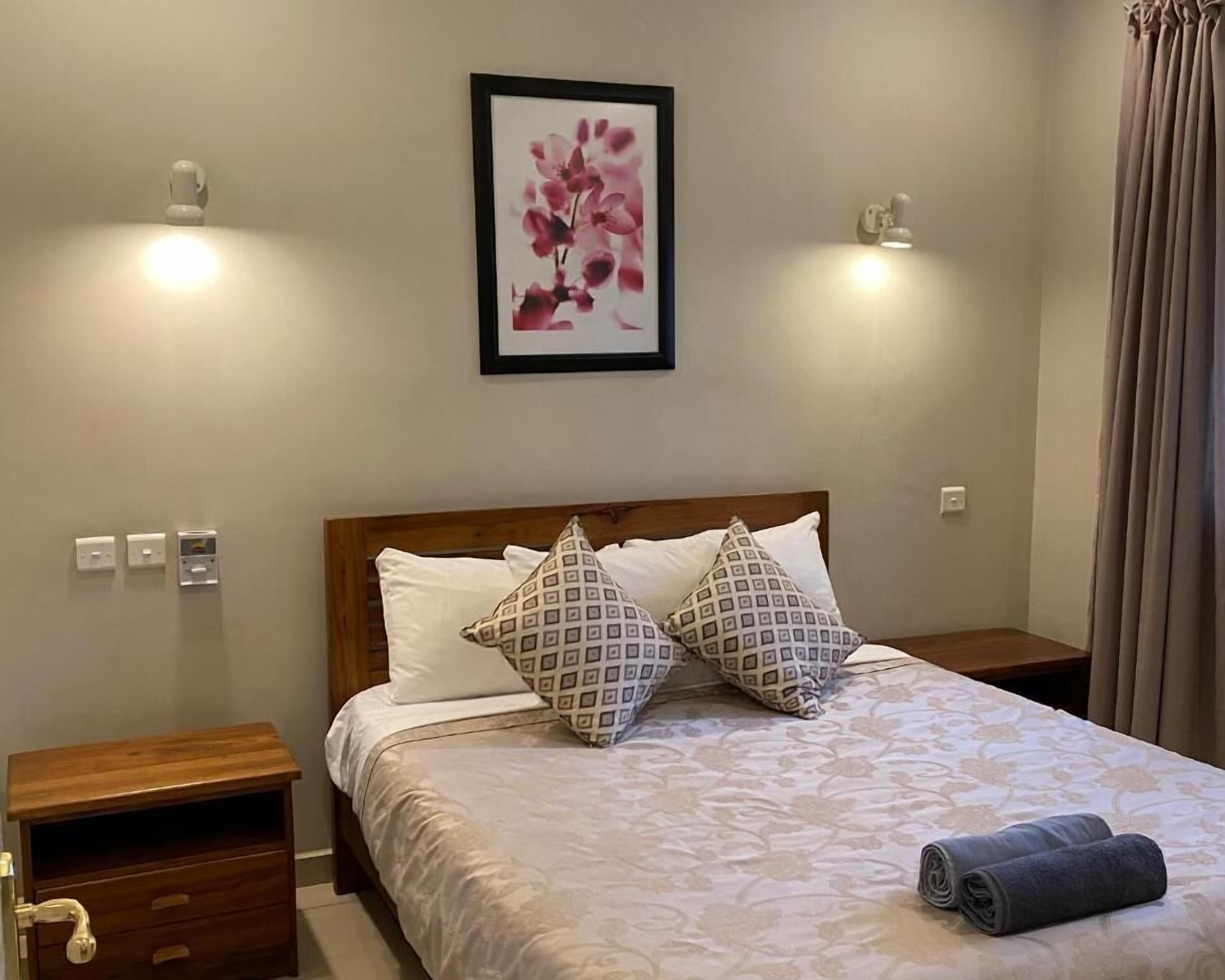 Una cama bien hecha con almohadas estampadas, una obra de arte floral enmarcada en la pared y una toalla enrollada sobre la cama.