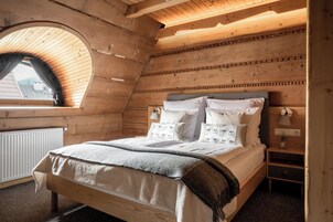A cozy attic bedroom with a wooden sloped ceiling, a large window, and a bed with multiple pillows and a blanket.