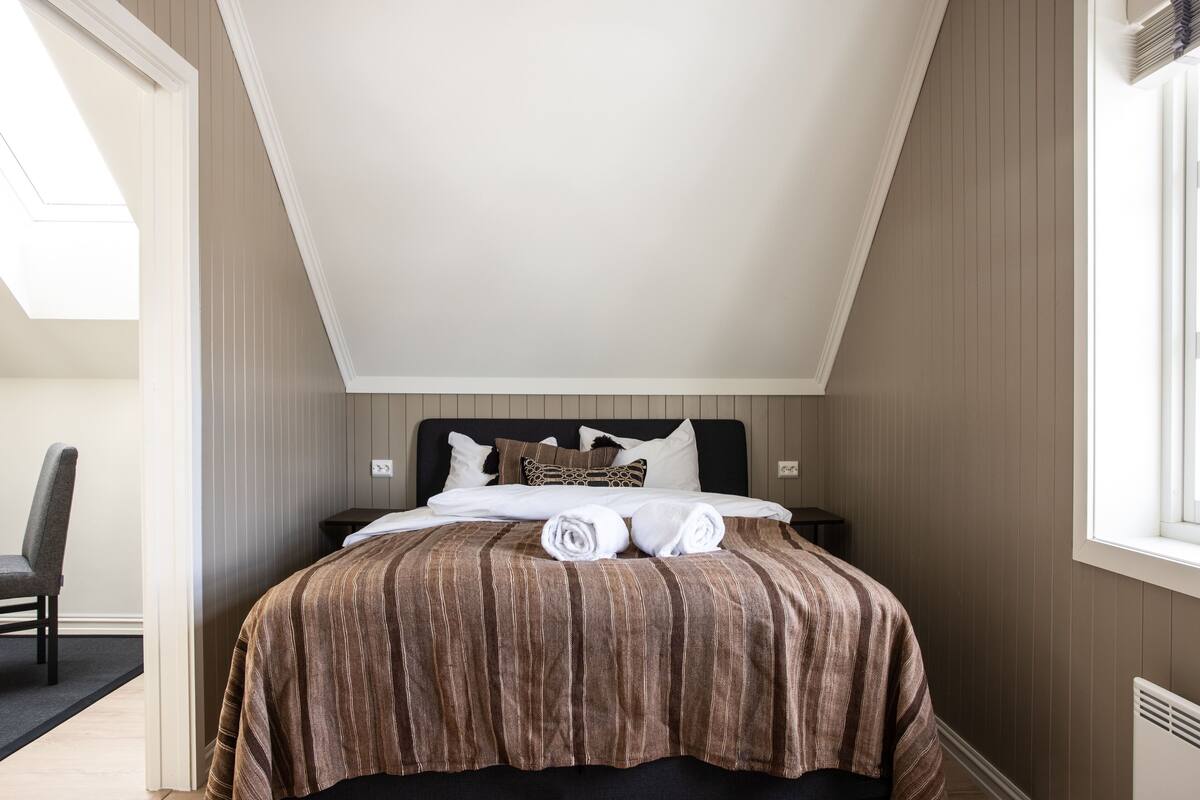 Une chambre avec un plafond en pente, un lit recouvert d’une courtepointe à rayures, des oreillers et une chaise.