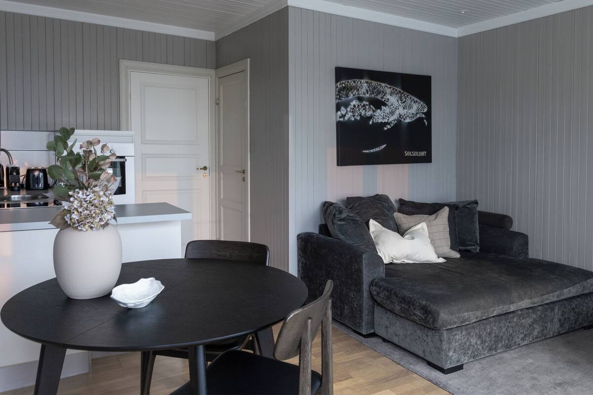 Un salon moderne avec une table ronde de couleur sombre, un canapé gris agrémenté de coussins, une porte blanche et une œuvre d’art encadrée accrochée au mur.