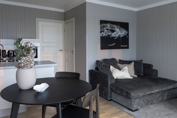 A modern living room with a dark round table, a grey sofa with cushions, a white door, and a framed artwork on the wall.