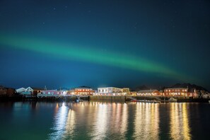 A waterfront hotel with a reflection of buildings in calm water, under a night sky with visible stars and the aurora borealis. - Solsiden Brygge (Vestvågøy)