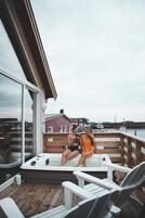 Una terraza de madera con jacuzzi y sillas de descanso, con vistas a un puerto deportivo con barcos y edificios.