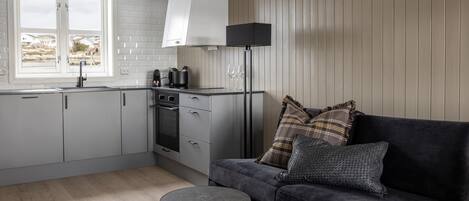 A modern kitchen with white cabinets, a black countertop, and a black floor lamp. There is a dark gray sofa with patterned cushions and a small round table.