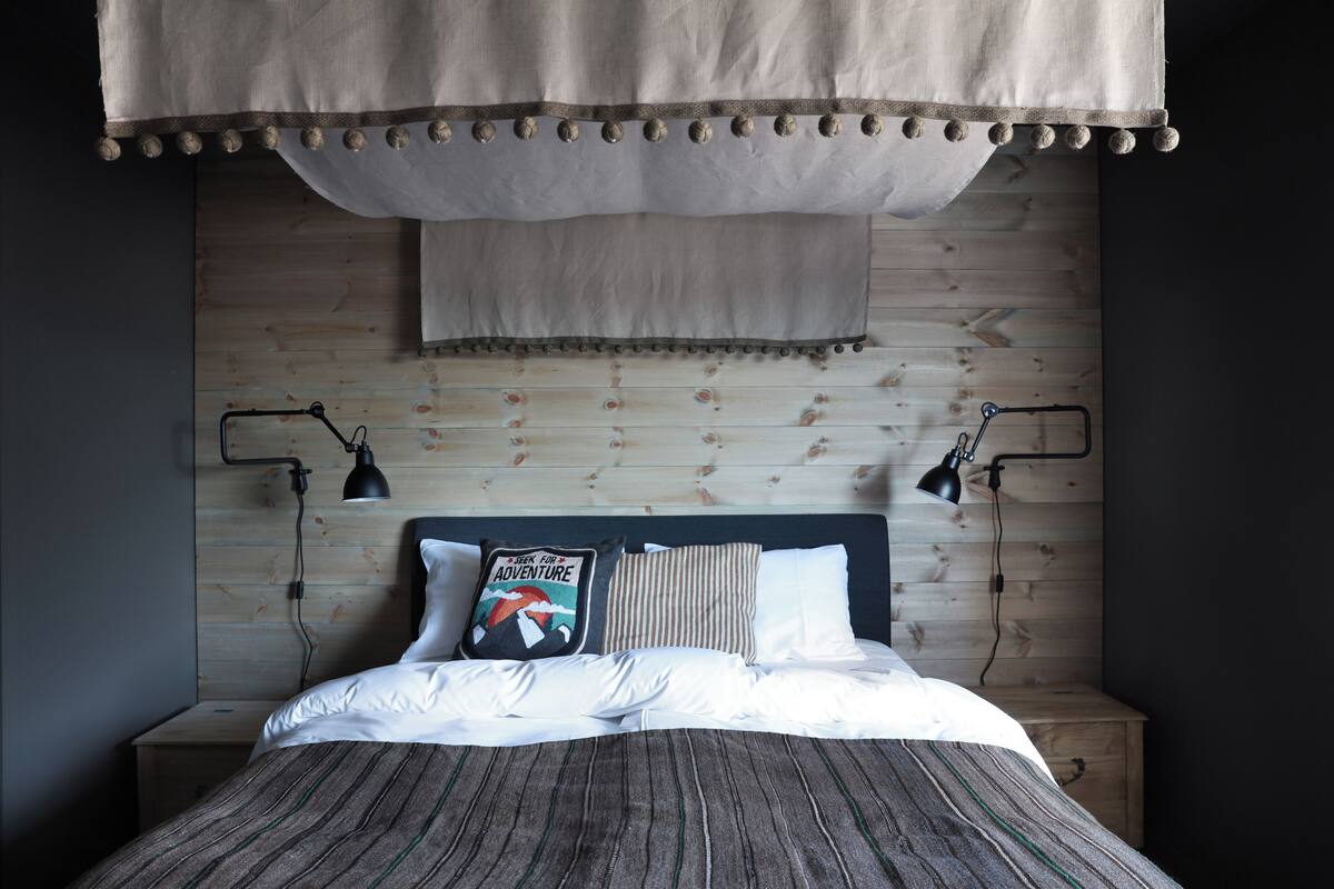 Un lit bien fait, avec une literie à rayures, un oreiller orné d’un motif sur le thème du voyage et une tête de lit adossée à un mur en bois.