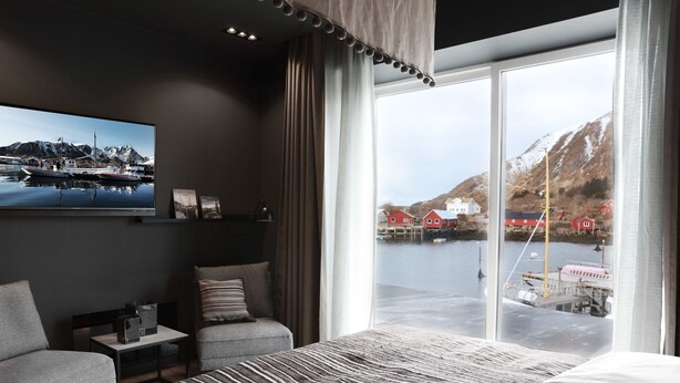 Une chambre d’hôtel moderne, dotée d’un grand lit, d’un bureau et d’une chaise. La chambre dispose d’une grande fenêtre offrant une vue sur un port et des montagnes.