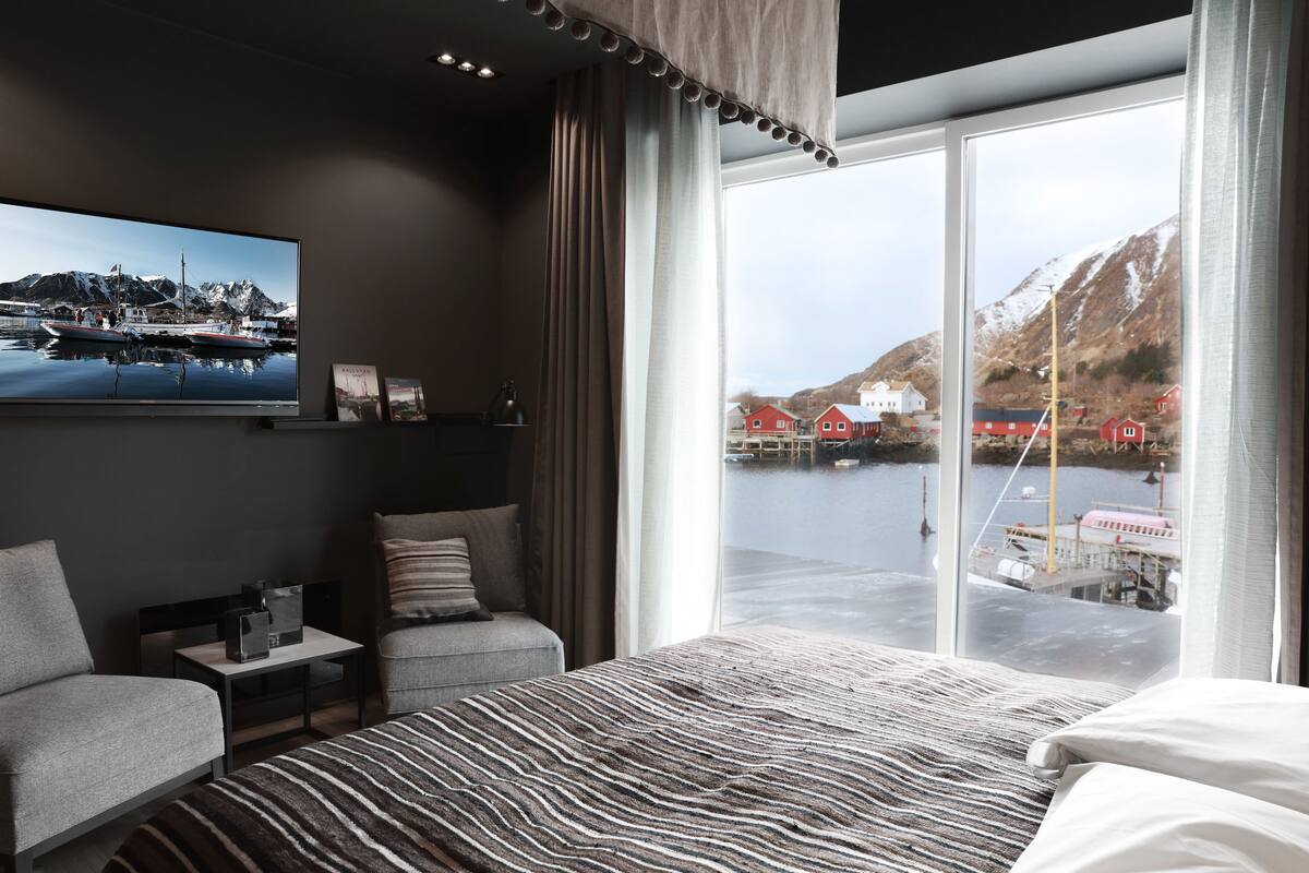 Une chambre d’hôtel moderne, dotée d’un grand lit, d’un bureau et d’une chaise. La chambre dispose d’une grande fenêtre offrant une vue sur un port et des montagnes.