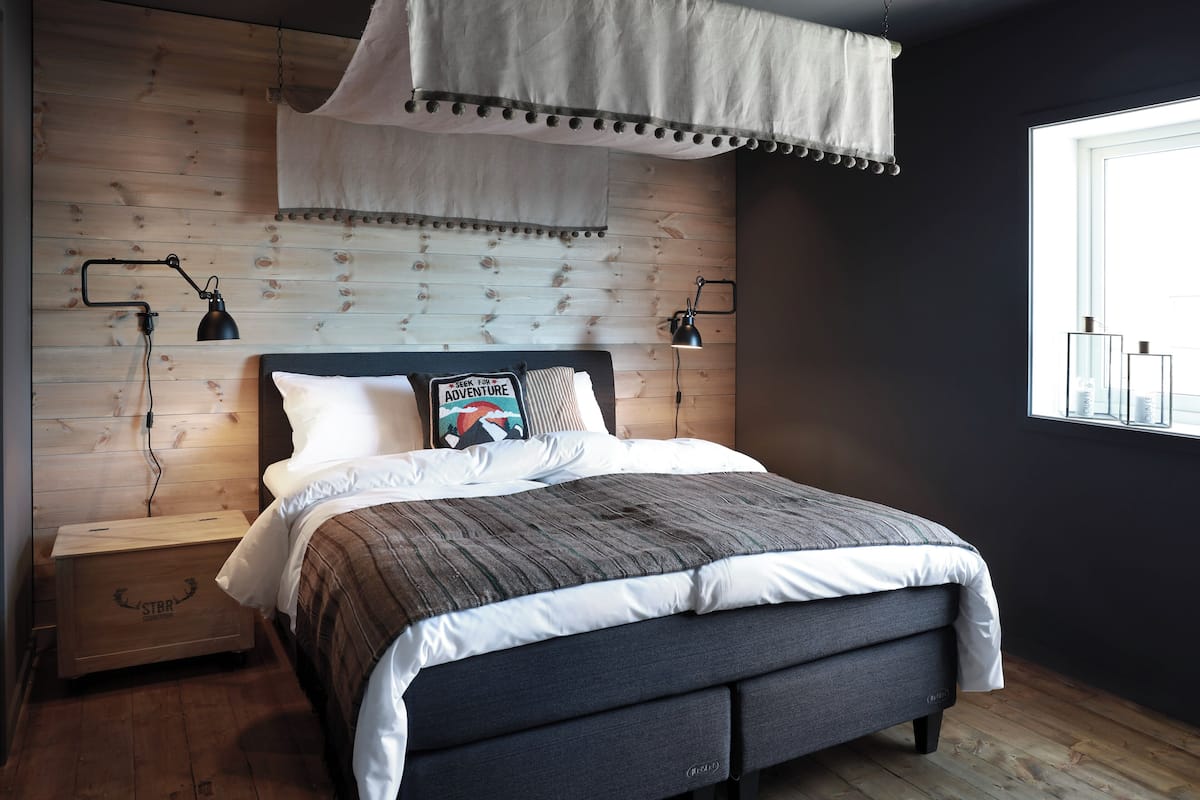 Une chambre moderne avec une tête de lit en bois, un lit recouvert d’une couverture à rayures, deux lampes de chevet et une fenêtre donnant sur des immeubles.