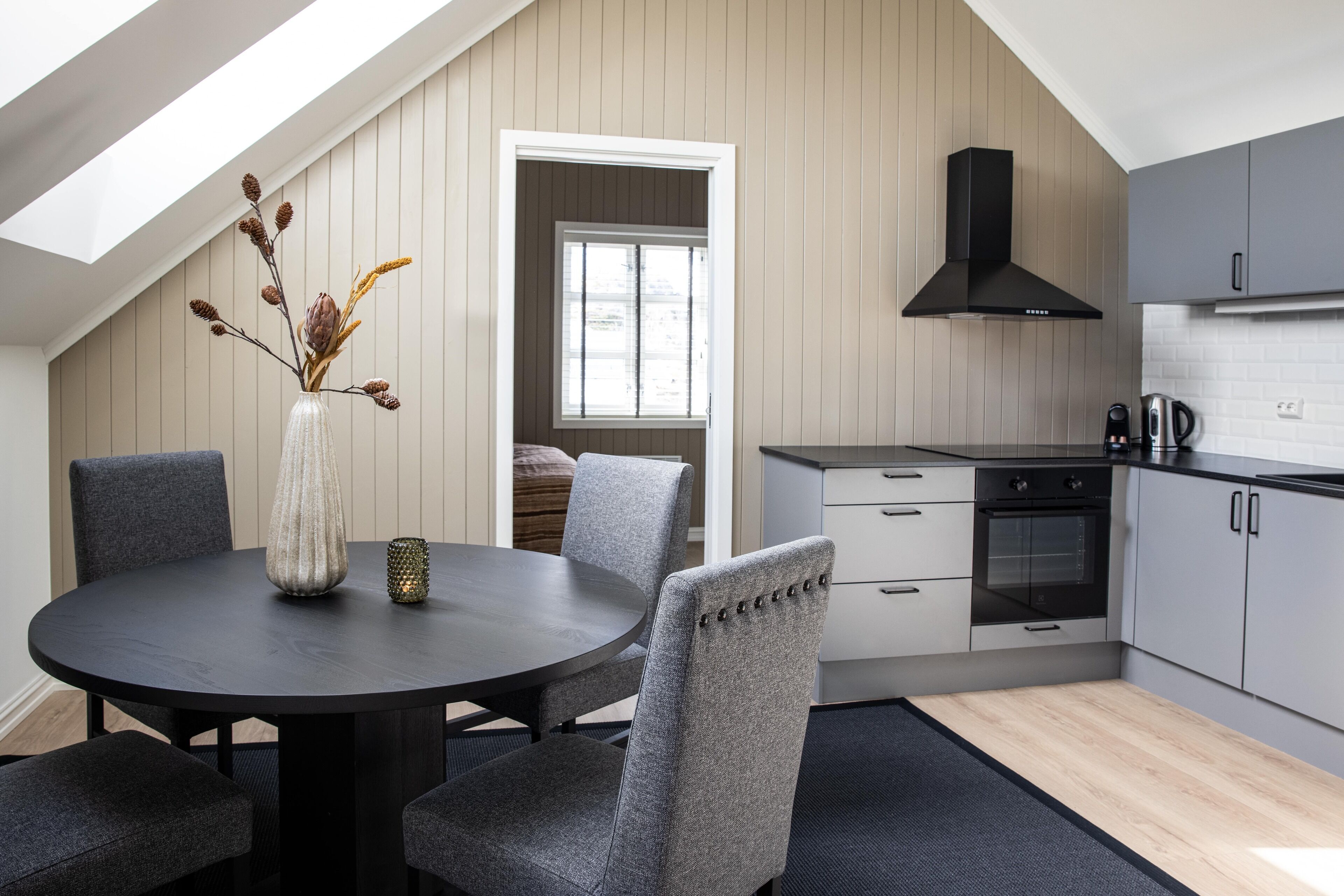 A modern kitchen with grey cabinetry, a black range hood, and a dining area with a round table and grey upholstered chairs.