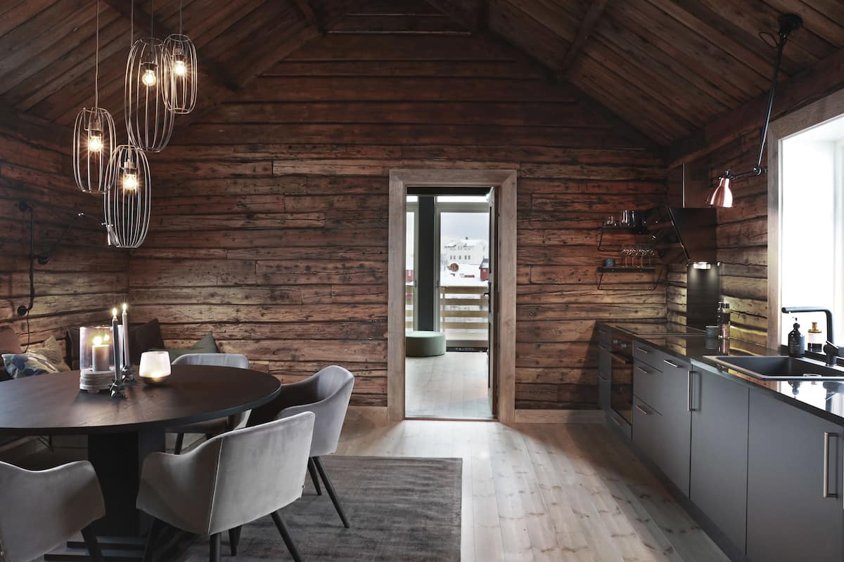 L’intérieur d’un chalet rustique comprend un coin repas en bois, une cuisine moderne et une vue sur le paysage, visible à travers une porte.