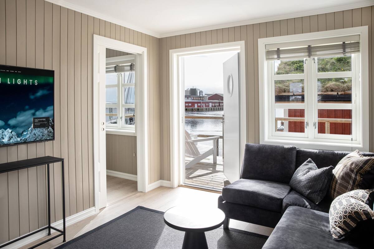 Un salon avec un canapé gris, une petite table basse ronde et un téléviseur fixé au mur. Deux portes donnent sur un balcon d’où l’on peut admirer une vue sur un quai et des bâtiments.