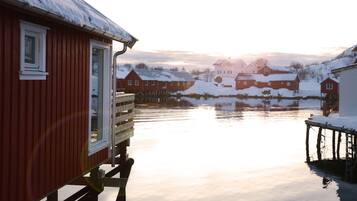 A red wooden building with a window and a door, adjacent to a calm water body, with snow-covered houses in the background.