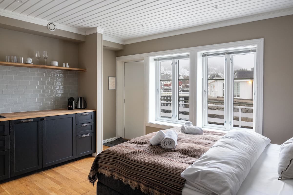 Une chambre moderne avec un grand lit, un coin cuisine doté d’un mur de fond en briques et une vue dégagée sur l’extérieur grâce à une grande fenêtre.