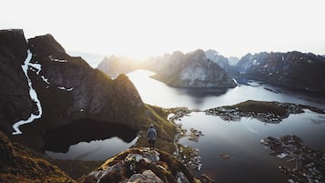 Un paisaje montañoso con una persona de pie sobre un afloramiento rocoso, contemplando un lago tranquilo y un pequeño asentamiento.