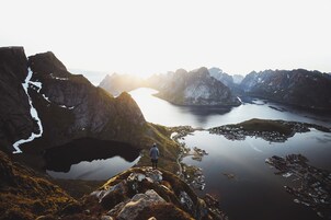 Un paisaje montañoso con una persona de pie sobre un afloramiento rocoso, contemplando un lago tranquilo y un pequeño asentamiento.