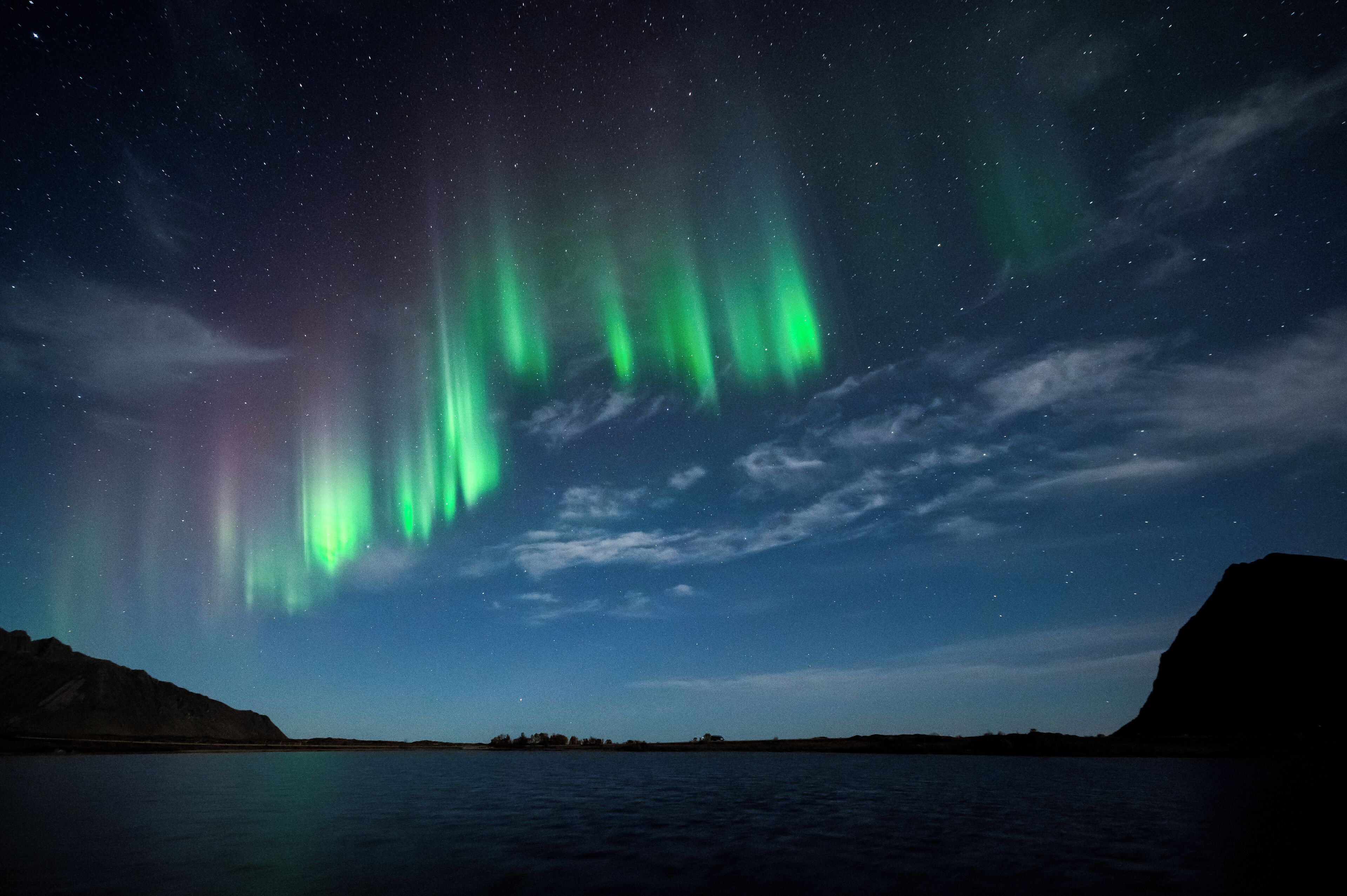 Céu noturno com auroras boreais refletidas em águas calmas e uma cadeia de montanhas ao longe.