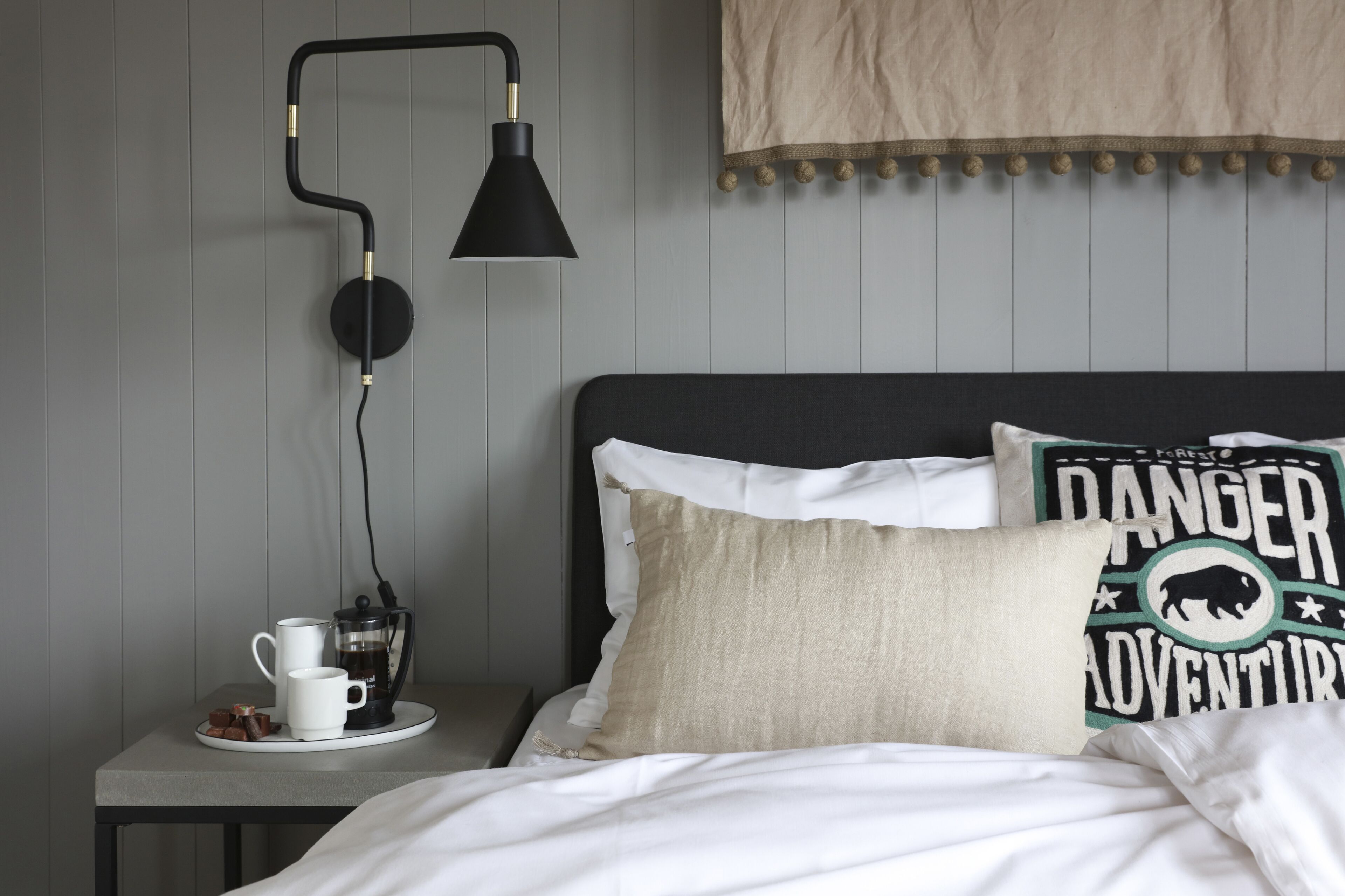 A neatly made bed with a white comforter and two decorative pillows, one with a 'Danger Adventure' design. A black lamp with a gold accent and a white mug with a coffee pot on a small table beside the bed.
