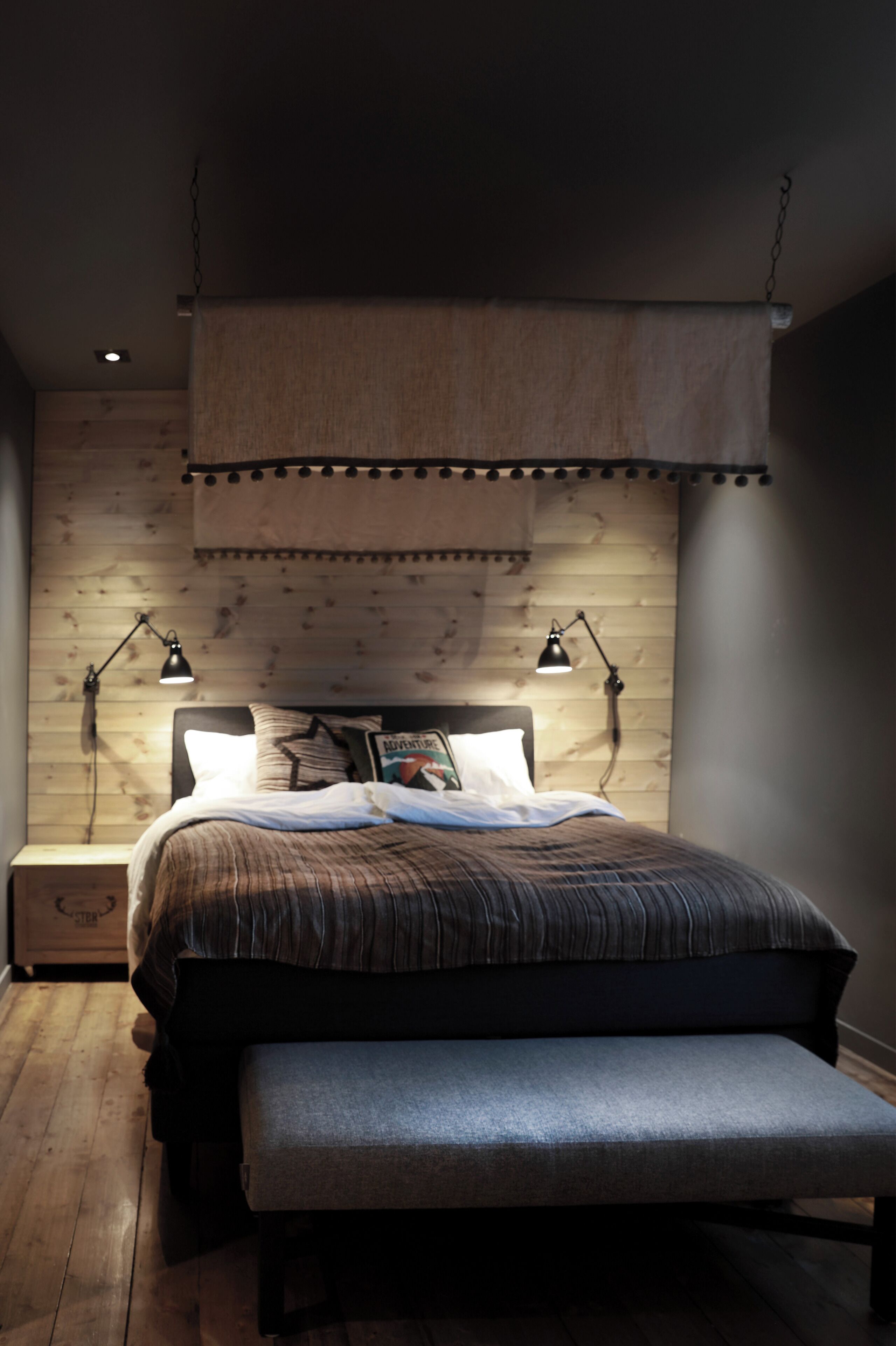 A bedroom with a wooden headboard, a bed with striped bedding, two bedside lamps, and a bench at the foot of the bed.