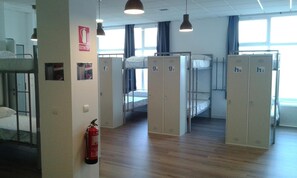 A dormitory room with bunk beds, lockers, a fire extinguisher, and a window with curtains.