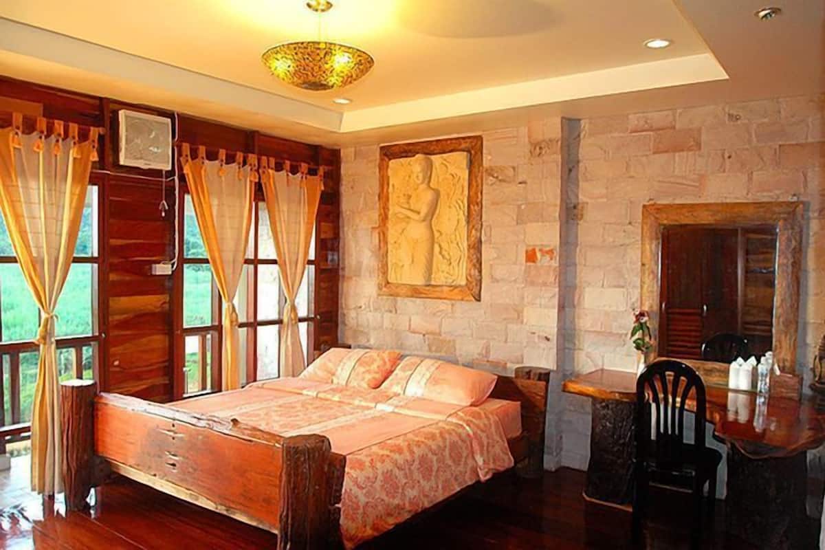 A bedroom with a wooden bed, a stone wall, a large window with curtains, and a small table with chairs.