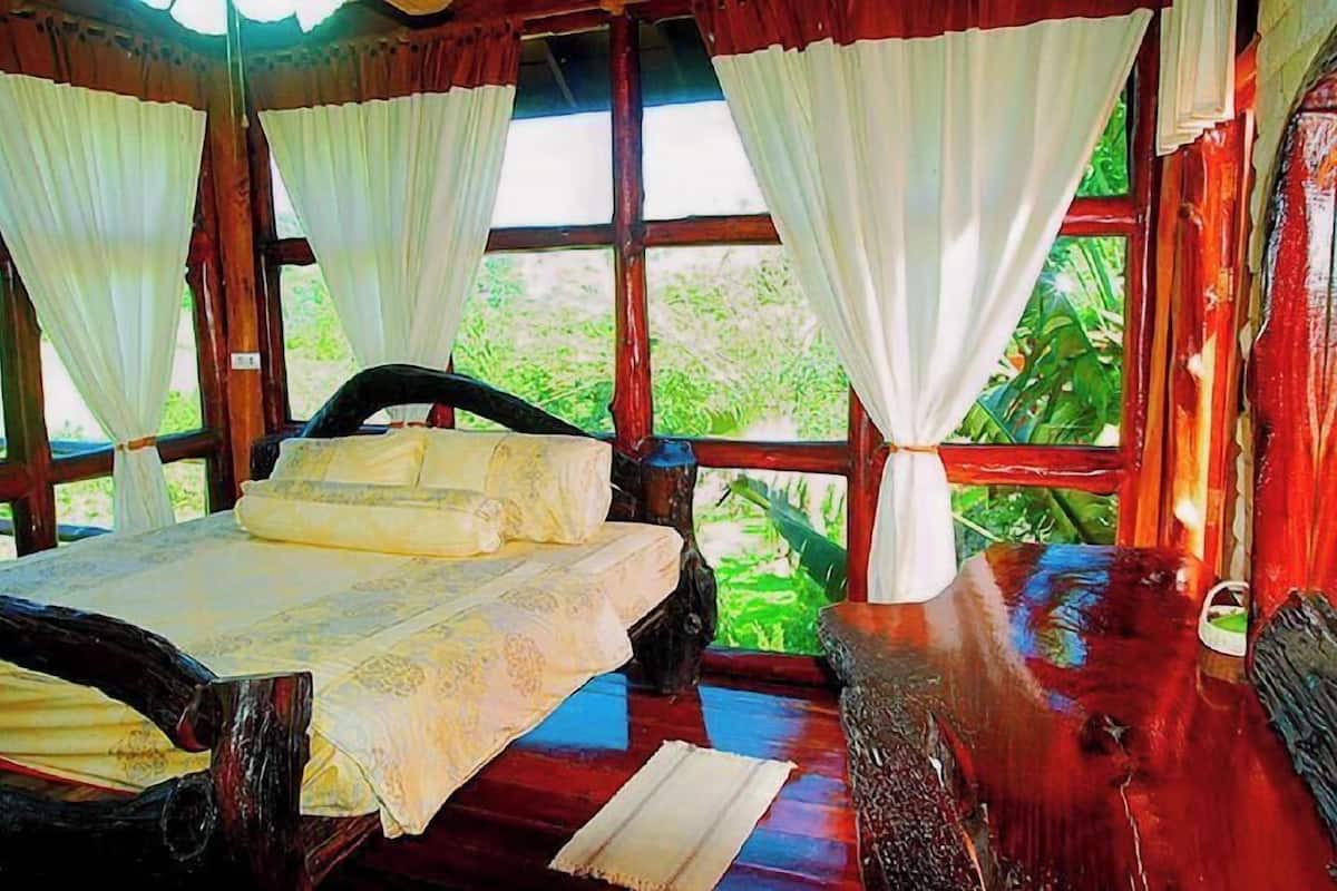 A wooden room with a bed, curtains, and a window with a view of greenery.