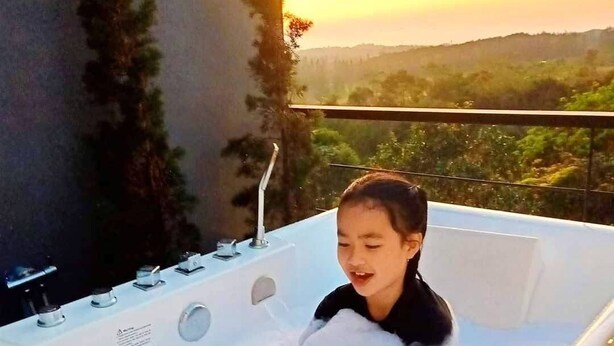 A person relaxing in a white bathtub with multiple faucets, set against a backdrop of a sunset and greenery. - Kirirath Resort (Pak Chong)