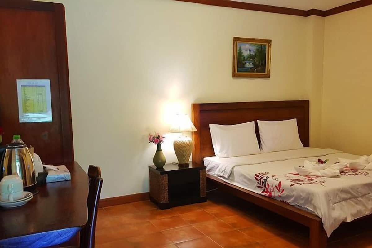 A hotel room with a bed, a nightstand with a lamp, a vase with flowers, a wooden desk with a chair, and a framed picture on the wall.